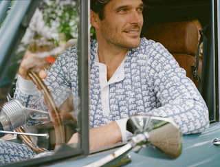 A man wearing a blue and white shirt with an elephant pattern sits in the driver’s seat of a vintage car, smiling and looking out the open window, capturing a sense of timeless style.