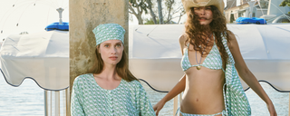 Two women in green and white patterned outfits pose by the water; one wears a headscarf and cover-up, the other a bikini, tote bag, and cowboy hat. Boats and trees are visible in the background.