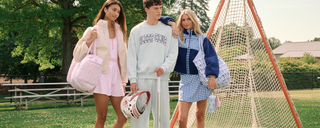 Three young adults in casual, pastel-colored outfits stand by a lacrosse goal on a sunny day. Two women carry bags, and a man holds a lacrosse helmet. Bleachers and trees are visible in the background.