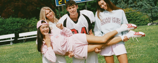 Three girls and one boy pose outdoors; the boy and two girls hold up the third girl, who is dressed in pink. They all look happy and playful. A scoreboard and benches are visible in the background.