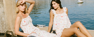 Two young women in floral print dresses sit by the water on a sunny day.