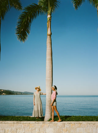 Two women standing next to a palm tree by the ocean with a clear blue sky.
