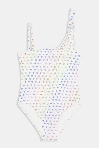 The Disco Hearts Kids Jacey One Piece by Roller Rabbit is a white swimsuit with rows of colorful hearts, featuring an asymmetrical neckline, one thin strap, and a knot-tie detail on the right shoulder.