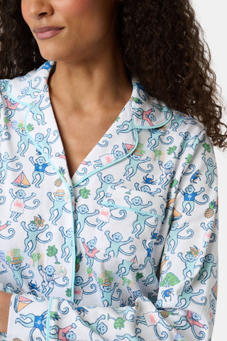 A woman with curly hair wears the Charleston Monkey Long Sleeve Polo Pajamas, featuring a white top with playful blue monkeys holding plants and sailboats, adding charm to any women’s sleepwear collection.