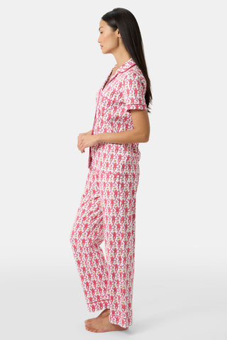 A woman stands barefoot in profile, wearing the Monkey Fiora Set—pink monkey print pajamas with short sleeves and long pants—against a plain light background.
