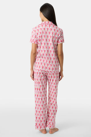 A woman with long dark hair stands barefoot, facing away, in the Monkey Fiora Set—matching pima cotton pajamas with short sleeves and long pants, featuring a playful animal print—against a plain light background.