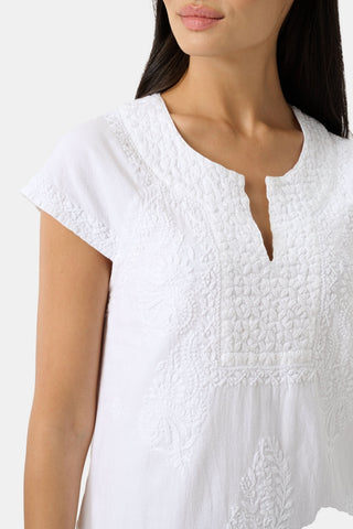 A woman with long, straight hair and a relaxed expression wears the Roller Rabbit Faith Dress—an elegant embroidered cotton dress with short sleeves and a knee-length hem—paired with green sandals against a plain light background.