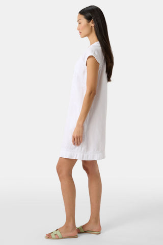 A woman with long, straight hair and a relaxed expression wears the Roller Rabbit Faith Dress—an elegant embroidered cotton dress with short sleeves and a knee-length hem—paired with green sandals against a plain light background.
