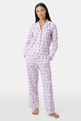 A woman stands barefoot in Palm Beach Monkey Long Sleeve Polo Pajamas, made from soft Pima cotton with a pastel floral and animal print. The collared top and loose pants evoke relaxed Palm Beach style against a plain light background.
