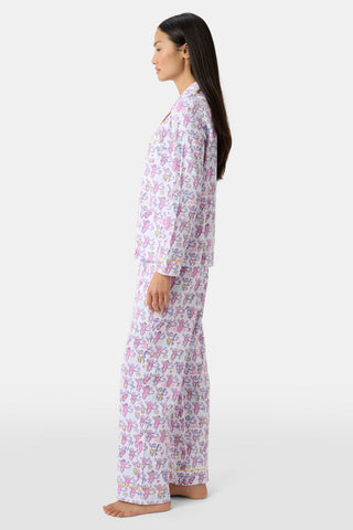 A woman with long dark hair stands in profile, barefoot against a white background, wearing Palm Beach Monkey Long Sleeve Polo Pajamas made from soft Pima cotton and featuring a colorful floral print.