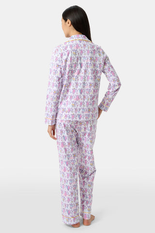 A woman with long dark hair stands barefoot, facing away, wearing Palm Beach Monkey Long Sleeve Polo Pajamas—classic Pima cotton with a white base and pink/purple floral print, evoking relaxed Palm Beach elegance.