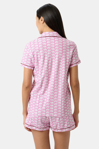 A woman with long dark hair faces away, wearing Hathi Polo Pajamas—a pink, Peruvian-made set with an elephant print, including a short-sleeve button-up top and ruffled shorts.