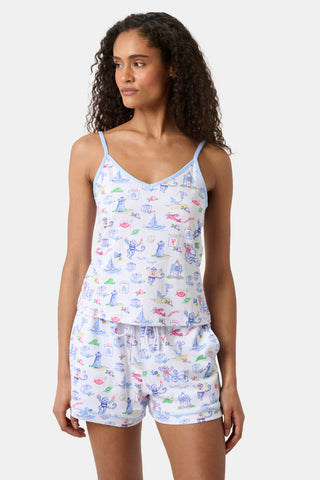 A woman wearing a white camisole and shorts pajama set with colorful cartoon prints stands facing forward, her left hand by her side, looking slightly to the right, against a plain light background. The playful cartoon motifs add a cheerful touch to the sleepwear ensemble.