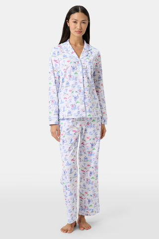 A woman stands barefoot wearing white button-up pajamas adorned with colorful, playful animal and object prints on both the top and pants. She faces forward against a plain, light background, showcasing whimsical designs that add a touch of fun to her sleepwear.