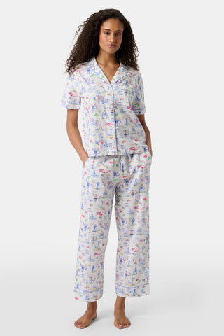 Woman with curly hair wearing a matching short-sleeve pajama set with a playful pastel animal print, standing barefoot against a white background. Hands are in her pockets as she looks forward.