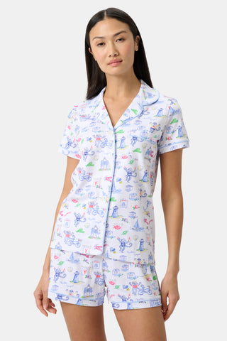 Woman wearing a short-sleeve button-up pajama top and matching shorts with playful blue, pink, and green cartoon prints, standing against a simple light background.