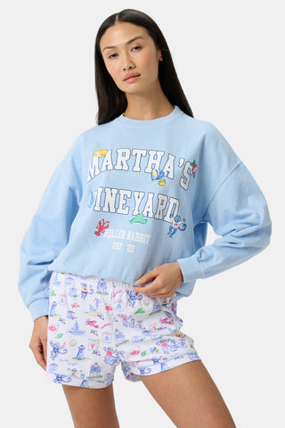A woman wears a light blue Martha's Vineyard sweatshirt with cartoon graphics and white shorts featuring colorful illustrations, standing against a plain light background.