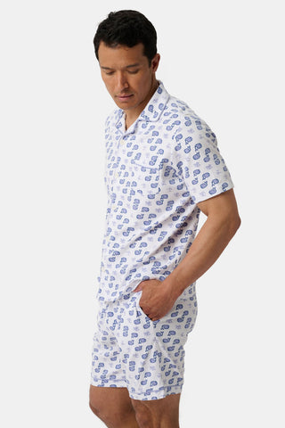A man stands with one hand in his pocket, wearing the Mens Moby Louie Pajamas—a matching white set featuring a blue printed pattern—against a plain light background.