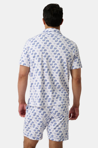 A man stands facing away, wearing the Mens Moby Louie Pajamas—a matching polo top and shorts set with a white base and repeating blue pattern—set against a plain, light background.