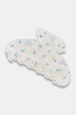 A close-up of dark hair secured with the Roller Rabbit Mini Monkey Blossom Claw Clip, a white, iridescent acrylic hair accessory adorned with small blue and green designs. The background is plain white.