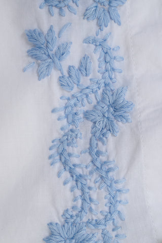 Close-up of the Vanna Embroidery Janna Cover Up by Roller Rabbit, showcasing light blue floral embroidery on white cotton voile. Delicate flowers and leaves add stylish detail—perfect as a chic beach cover up.