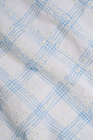 The Sun Check Eyelet Aurora One Piece by Roller Rabbit features white fabric with a light blue plaid pattern and white embroidered florals, giving it a textured, delicate look similar to soft, slightly wrinkled cloth.