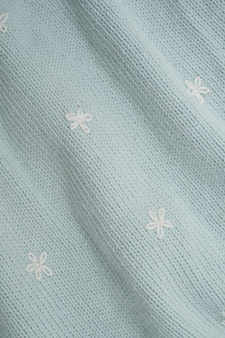 A woman with curly hair wears the Roller Rabbit Floral Bloom Nyla Cardigan, a light blue short-sleeved top with small white flowers, paired with loose white embroidered pants, standing against a plain light background.