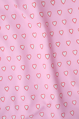 A young girl with long brown hair smiles as she wears the Roller Rabbit Kids Hearts Dory One Piece, a sleeveless pink dress with a flared, ruffled skirt and heart print, standing with her hands behind her back against a light background.