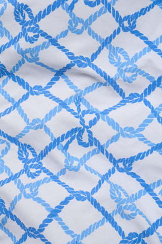 A young girl with long brown hair wears the Roller Rabbit Kids Seaside Knots Jacey One Piece, featuring a blue geometric pattern and a bow on one shoulder, smiling with her hands on her hips against a plain background.