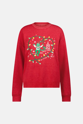 A smiling woman sits on a wooden bench wearing a red Deck The Halls Andie Sweatshirt featuring an illustrated monkey in string lights and patterned Bea Sweatpants.