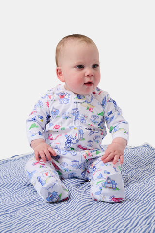 A baby wearing a white onesie with colorful drawings sits on a blue and white striped blanket, gazing curiously to the side.