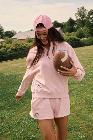 A young woman in a pink sweatshirt and Roller Rabbit Game Day Nicki Shorts smiles while holding a football in one hand, standing on a grassy field with trees and buildings in the background.