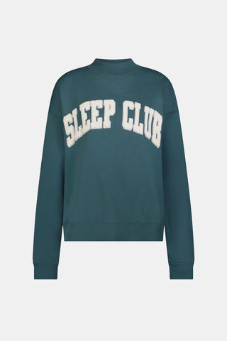A woman wears an ARILCONFEX Sleep Club Taylor Mockneck in green with a long pale pink scarf and white shorts featuring a small red pattern.