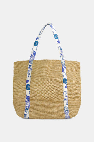 A woman in a sleeveless white eyelet dress carries the Amanda Woven Beach Bag by Roller Rabbit, featuring blue and white patterned shoulder straps. The large bag is shown from the side as she walks.