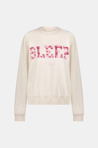 A woman wears the ARILCONFEX Sleep Club Andie Sweatshirt, an oatmeal crewneck with SLEEP in pink plaid appliqué across the chest and Club embroidered below it in pink script.