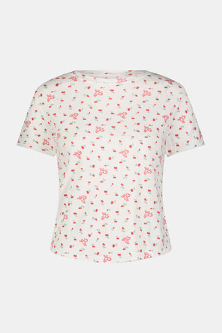 A smiling woman with curly hair stands outdoors near a window, wearing the Roller Rabbit Mini Monkey Blossom Pointelle Classic Tee—a white short-sleeve top featuring a delicate pink floral print.