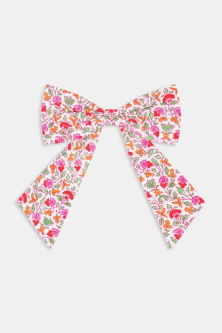 The Garden Gate Floral Hair Bow by Roller Rabbit is a large cotton bow with long tails, showcasing a vibrant pattern of pink, red, and orange flowers with green leaves on a white background.
