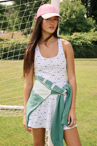The Roller Rabbit Tennis Hearts Becky Active Dress is a sleeveless white active dress with a V-neckline and a flared skirt, adorned with an all-over pattern of small blue and green heart shapes. The dress is shown against a plain light gray background.