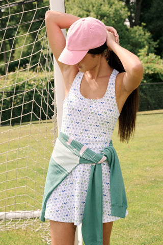 The Roller Rabbit Tennis Hearts Becky Active Dress is a sleeveless white active dress with a V-neckline and a flared skirt, adorned with an all-over pattern of small blue and green heart shapes. The dress is shown against a plain light gray background.