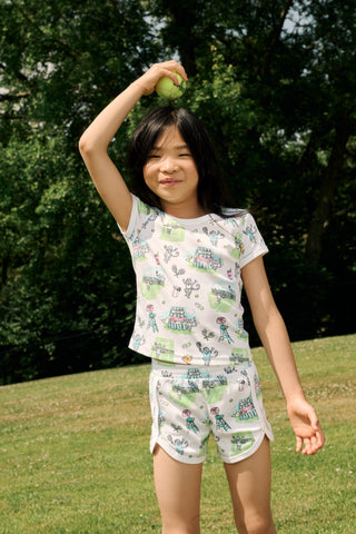 A girl stands outdoors in a Roller Rabbit Kids Game, Set, Monkey Playa Play Set, holding out a tennis ball toward the camera. Her playful outfit is perfect for kids’ tennis activities, and the photo is taken from a low angle with blue sky and trees in the background.