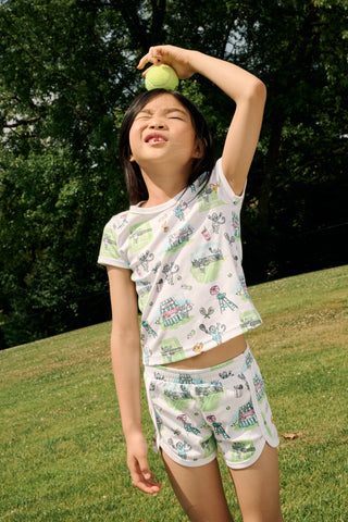 A girl stands outdoors in a Roller Rabbit Kids Game, Set, Monkey Playa Play Set, holding out a tennis ball toward the camera. Her playful outfit is perfect for kids’ tennis activities, and the photo is taken from a low angle with blue sky and trees in the background.