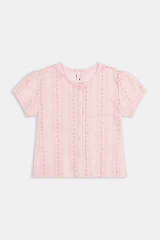 A young girl in a rose pink dress smiles while kneeling on the floor, holding a colorful folder. A pink backpack hangs nearby in a classroom or locker area, and she wears her cozy Roller Rabbit Kids Daisy Heart Stripe Diana Cardigan.