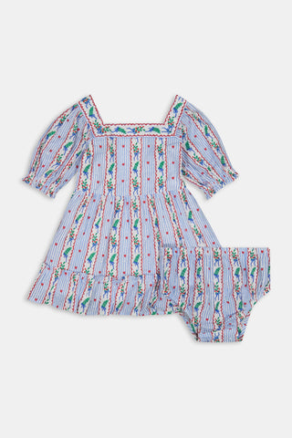 A girl wears the SANSKRITI Kids Mistletoe Stripe Lottie Dress in a blue and white-striped cotton print with puff sleeves, a square neckline, and red heart accents, paired with blue velvet Mary Jane flats.