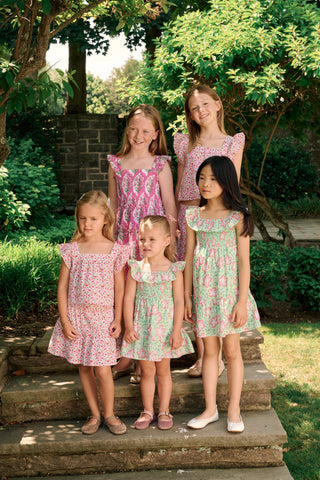 The Kids Verdant Floral Ryan Dress by Roller Rabbit is a green dress for little ones, showcasing pink and white hibiscus flowers in a floral block print. It features ruffled sleeves, a matching diaper cover, and is crafted from soft cotton for comfortable pullover wear.