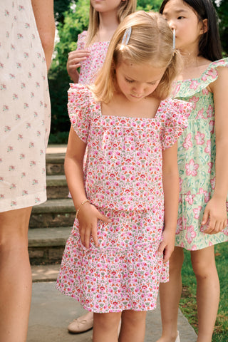 Three young girls sit close together outdoors, each wearing the Roller Rabbit Kids Garden Gate Floral Emmett Top—an ideal choice for girls' summer clothing. They are smiling and appear to be engaged in conversation, with greenery and stone walls in the background.