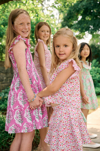 The Kids Ava Floral Ceri Dress by Roller Rabbit is a sleeveless baby dress featuring a smocked bodice and ruffled straps, adorned with green and white leaf patterns on a vibrant pink background, and comes with matching bloomers.