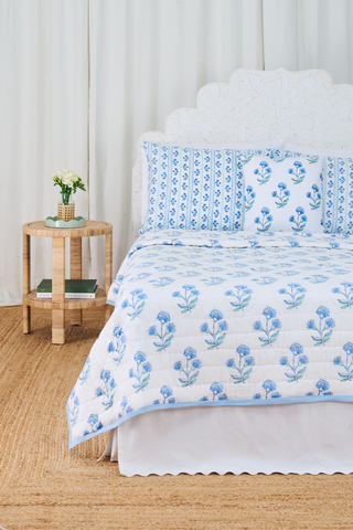 A neatly made bed with the Roller Rabbit Rosa Ditsy Floral Quilt sits beside a round wicker nightstand with a vase of white flowers and stacked books, atop a woven rug and framed by white curtains in the background.