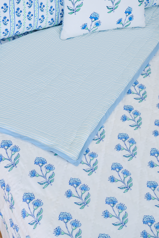 A neatly made bed with the Roller Rabbit Rosa Ditsy Floral Quilt sits beside a round wicker nightstand with a vase of white flowers and stacked books, atop a woven rug and framed by white curtains in the background.