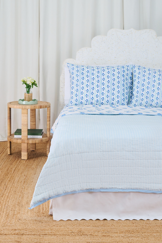 A neatly made bed with the Roller Rabbit Rosa Ditsy Floral Quilt sits beside a round wicker nightstand with a vase of white flowers and stacked books, atop a woven rug and framed by white curtains in the background.