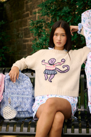 A young woman sits on a bench, wearing the Roller Rabbit Game Day Brittany Sweater with a cartoon figure, layered over a light dress. She is beside a blue patterned backpack and surrounded by greenery.